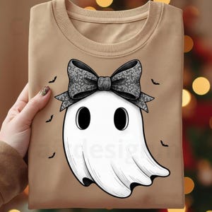 Op de afbeelding: Een beige sweatshirt met een wit spookontwerp met een patroon strik en zwarte ogen. Kleine zwarte vleermuissilhouetten zijn verspreid rond het spook. Het ontwerp is gecentreerd op de voorkant.