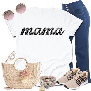 以下が含まれることがあります： 白のTシャツに「mama」の文字が白黒のヒョウ柄でプリントされています。画像には、ハイウエストのブルージーンズ、ヒョウ柄のアクセントが付いたベージュのスニーカー、麦わらハンドバッグ、サングラス、腕時計、ブレスレットも含まれています。