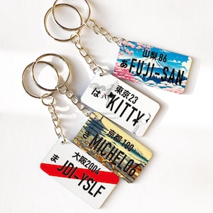 Peut inclure: Quatre porte-clés avec des étiquettes rectangulaires à motifs japonais. L'une affiche "FUJI-SAN" avec une scène de montagne, une autre "KITTY" avec une grue, et deux autres ont du texte japonais et des motifs de plaques d'immatriculation. Chaque porte-clés a une chaîne argentée et un anneau doré.