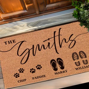 May include: A brown coir doormat with the text "The Smiths" in a cursive font. The doormat also features footprints, paw prints, and flip-flops with the names "William", "Marry", "Paddie", and "Chip" underneath.