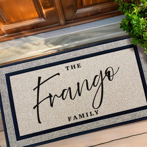May include: A rectangular doormat with the words "THE Frango FAMILY" in black script. The mat has a light gray textured center, a dark blue border, and a gray outer edge. The mat is placed in front of a wooden door.