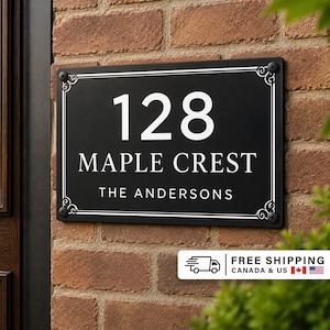 May include: A rectangular black house sign with white text featuring the number 128, the words MAPLE CREST, and the name THE ANDERSONS. The sign has a white border with decorative corner flourishes and is mounted with four black screws.