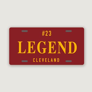 Puede incluir: Una placa de matr&iacute;cula rectangular de color granate con el texto amarillo #23, LEGEND y CLEVELAND. La placa tiene cuatro ranuras de montaje de color gris oscuro situadas cerca de las esquinas, con un dise&ntilde;o limpio y letras de alto contraste.