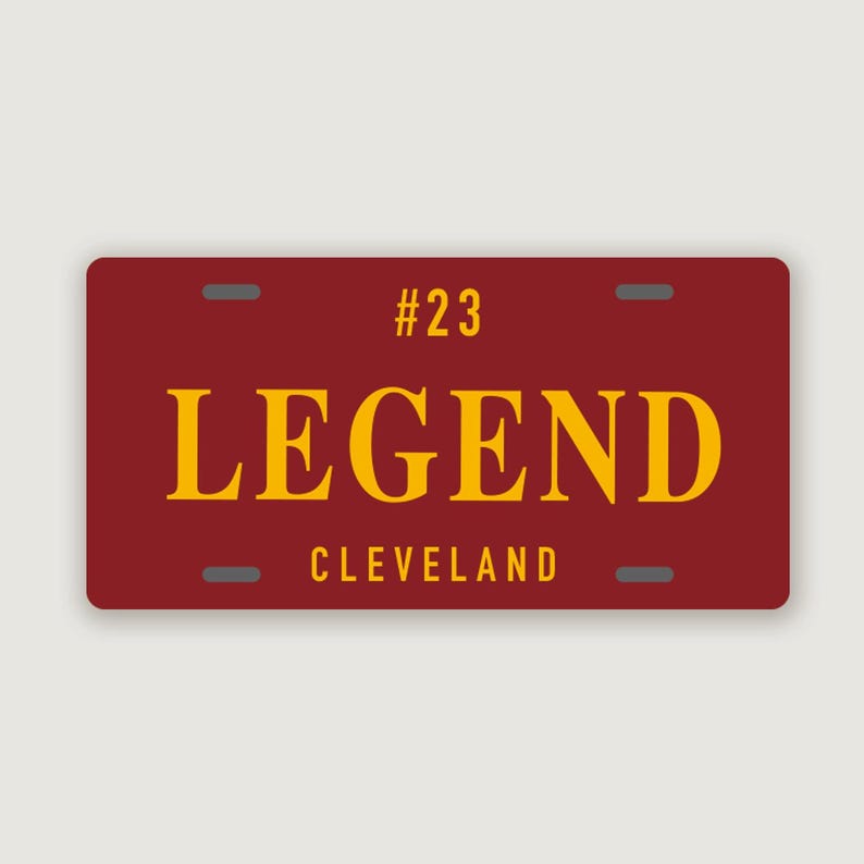 Puede incluir: Una placa de matr&iacute;cula rectangular de color granate con el texto amarillo #23, LEGEND y CLEVELAND. La placa tiene cuatro ranuras de montaje de color gris oscuro situadas cerca de las esquinas, con un dise&ntilde;o limpio y letras de alto contraste.
