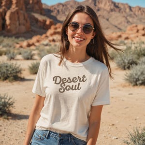 以下が含まれることがあります： 「Desert Soul」の文字がダークブラウンの筆記体で書かれた、クリーム色のTシャツ。青いジーンズと合わせています。背景には、山とまばらな植生のある砂漠の風景が広がっています。
