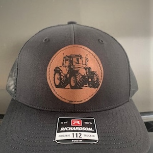 May include: Black and brown youth trucker hat with a brown leather patch featuring a tractor silhouette. The patch has the text "EST. 1970 RICHARDSON. ORIGINAL 112 TRUCKER YOUTH".