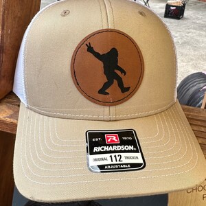 May include: A tan and white trucker hat with a brown leather patch featuring a black silhouette of a Bigfoot figure making a peace sign. The hat has an adjustable strap and a label that reads "Richardson. Original 112 Trucker Adjustable."
