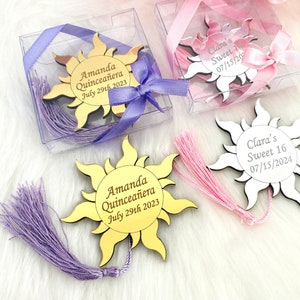 May include: Two gold and white sun-shaped mirror acrylic bookmarks with tassels. The gold bookmark has the text "Amanda Quincea&ntilde;era July 29th 2023". The white bookmark has the text "Clara's Sweet 16 07/15/2024".