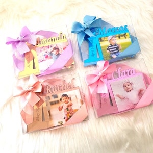 May include: Four personalized first birthday photo frames with a clear plastic box, a bow, and a photo of a baby. The frames are decorated with the names Amanda, Rachel, Marcos, and Clara, and the dates of the babies' birthdays.