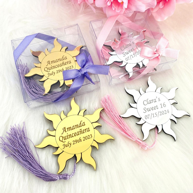 May include: Two gold and white sun-shaped mirror acrylic bookmarks with tassels. The gold bookmark has the text "Amanda Quincea&ntilde;era July 29th 2023". The white bookmark has the text "Clara's Sweet 16 07/15/2024".
