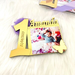May include: Gold-colored wooden frame with a photo of a baby girl, the number one, the name Amanda, and the date 11/08/2023. A gold butterfly is attached to the frame.