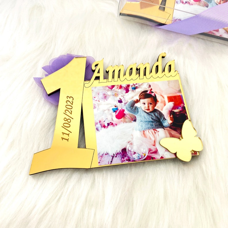May include: Gold-colored wooden frame with a photo of a baby girl, the number one, the name Amanda, and the date 11/08/2023. A gold butterfly is attached to the frame.