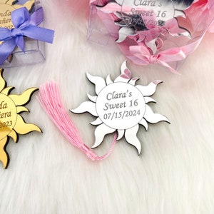 May include: A silver mirror sun with a pink tassel, engraved with "Clara's Sweet 16 07/15/2024".