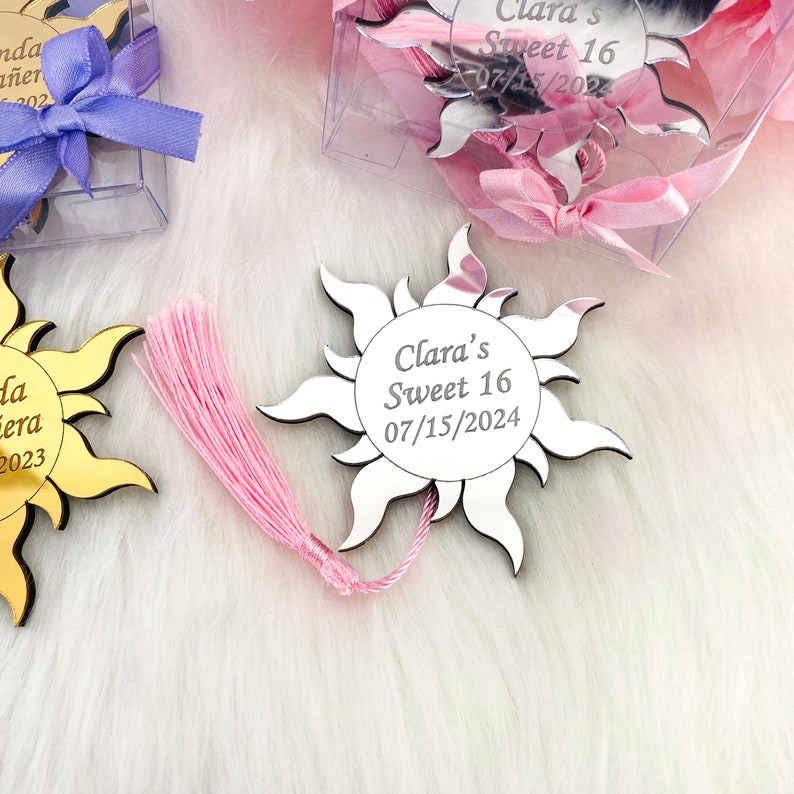 May include: A silver mirror sun with a pink tassel, engraved with "Clara's Sweet 16 07/15/2024".