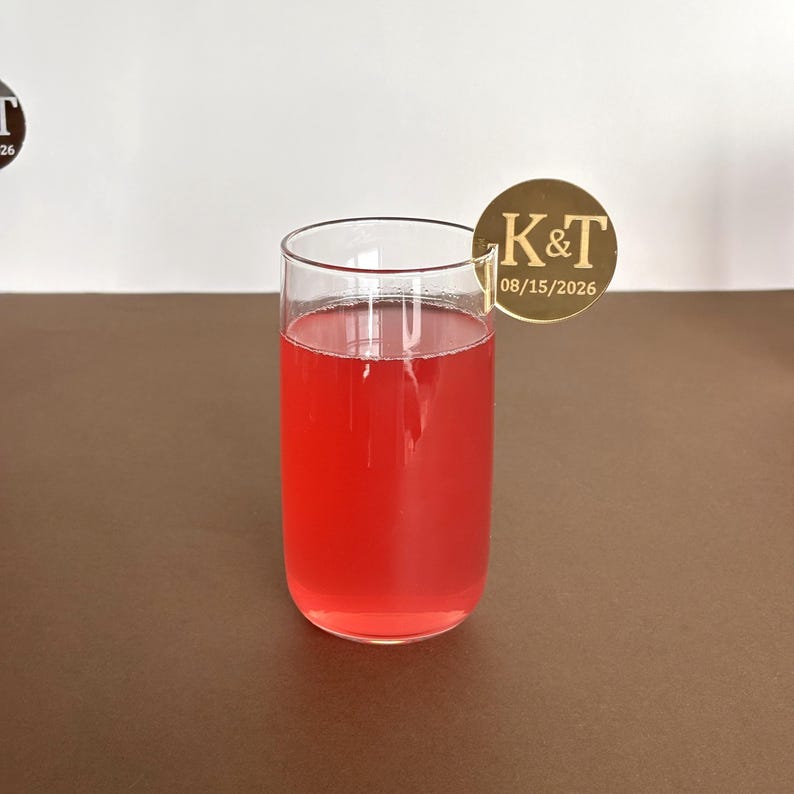 May include: A tall, clear glass filled with a vibrant red liquid, possibly a cocktail or juice. A gold-colored circular drink charm with the initials "K&T" and the date "08/15/2026" is attached to the rim. The drink sits on a brown surface.