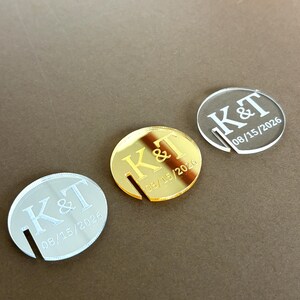 May include: Three acrylic wedding favors in silver, gold, and clear. Each disc features the initials "K&T" and the date "08/15/2026" engraved in a serif font. A slot is cut into the side of each disc.