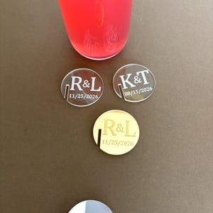 May include: Three personalized acrylic drink coasters. Two are clear with the initials "R&L" and "K&T" and dates. The third coaster is gold with the initials "R&L" and a date. A glass of red liquid is in the background.