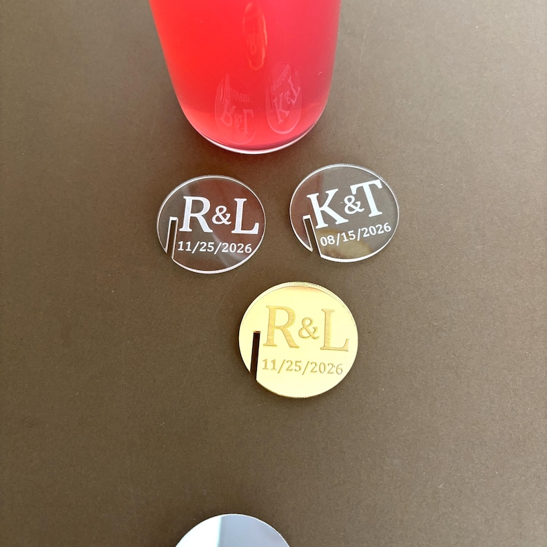 May include: Three personalized acrylic drink coasters. Two are clear with the initials "R&L" and "K&T" and dates. The third coaster is gold with the initials "R&L" and a date. A glass of red liquid is in the background.