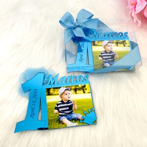 May include: Blue and white photo frame with a picture of a baby boy. The frame is decorated with a blue bow and a crown. The frame says "Marcos" and "April 1, 2023".