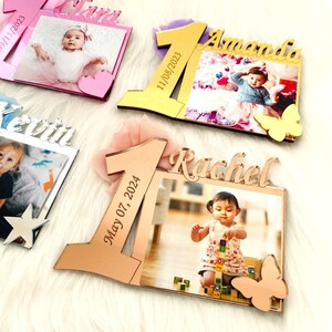 May include: Four personalized photo frames with a number one design, each with a different name and date. The frames are made of mirrored acrylic and have a gold, rose gold, silver, and pink finish. Each frame includes a butterfly accent.