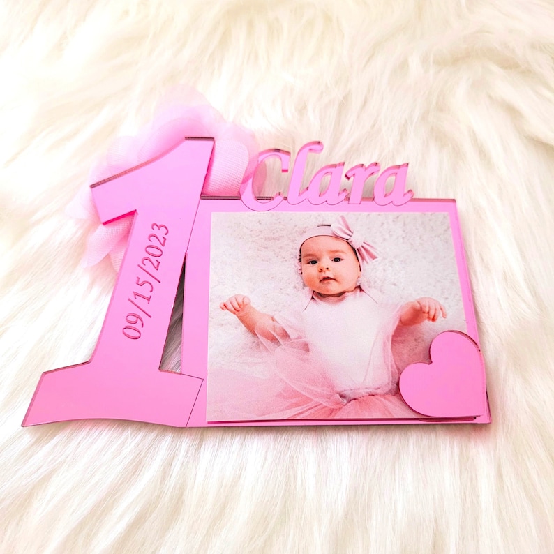 May include: Pink acrylic photo frame with a large number one and the name Clara. The frame has a space for a photo and the date 09/15/2023 is engraved on the number one.