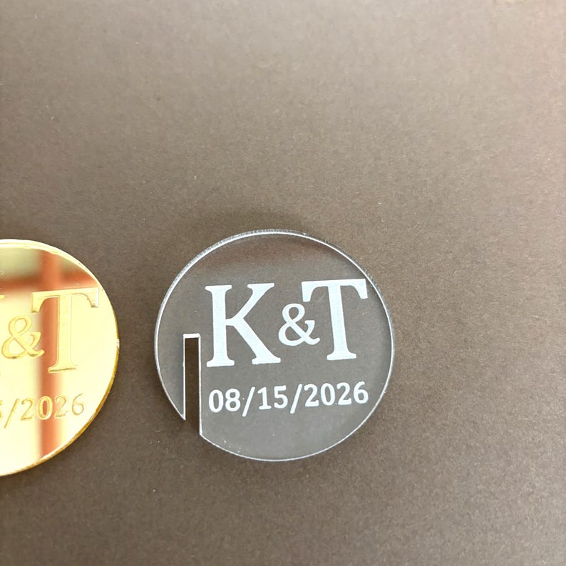 May include: Two circular wedding favors. One is gold with the letters "T" and "&" and the number "/2026". The other is clear with the letters "K&T" and the date "08/15/2026" in white.