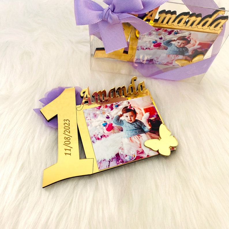 May include: Gold-colored wooden frame with a photo of a baby girl and the name "Amanda" written on it. The frame is shaped like the number one and includes the date "11/08/2023".