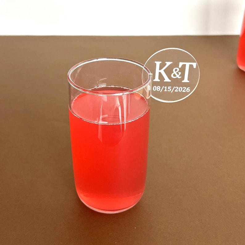 May include: A clear glass filled with a vibrant red liquid. The glass has a simple, cylindrical shape. A white circular sticker with the text "K&T" and the date "08/15/2026" is attached to the glass. The drink is set against a brown background.