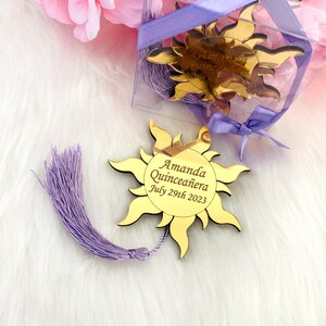 May include: Gold sun-shaped mirror with a purple tassel. The mirror has the text "Amanda Quincea&ntilde;era July 29th 2023" engraved on it.