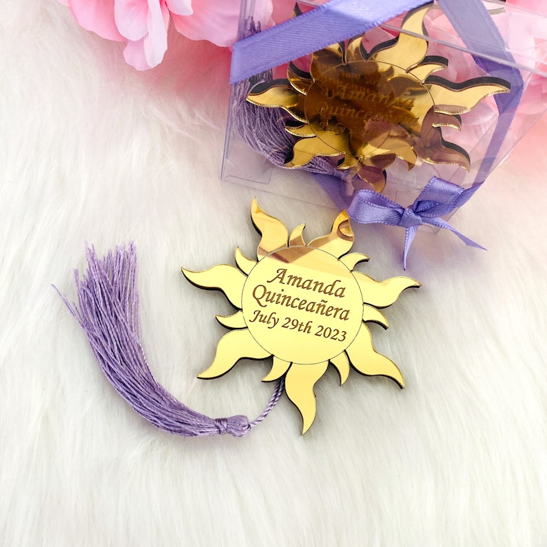 May include: Gold sun-shaped mirror with a purple tassel. The mirror has the text "Amanda Quincea&ntilde;era July 29th 2023" engraved on it.