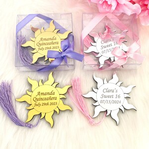 May include: Two sets of gold and white sun-shaped acrylic bookmarks with tassels. The gold bookmark has the text "Amanda Quincea&ntilde;era July 29th 2023" and the white bookmark has the text "Clara's Sweet 16 07/15/2024".
