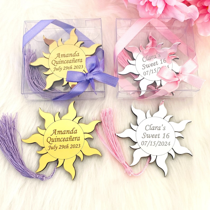 May include: Two sets of gold and white sun-shaped acrylic bookmarks with tassels. The gold bookmark has the text "Amanda Quincea&ntilde;era July 29th 2023" and the white bookmark has the text "Clara's Sweet 16 07/15/2024".