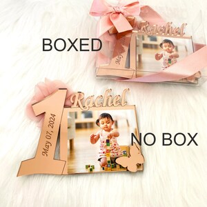 May include: Rose gold 1st birthday photo frame with a pink bow and the name Rachel and the date May 07, 2024. The frame has a picture of a baby girl playing with blocks.