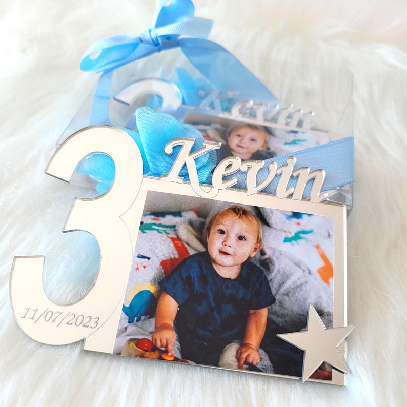 May include: A silver photo frame with a picture of a young boy. The frame is shaped like the number 3 and has the name "Kevin" and the date "11/07/2023" engraved on it. A blue ribbon is tied around the frame.