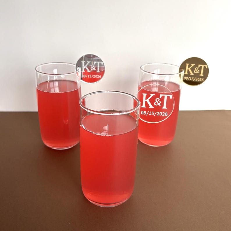 May include: Three clear glass tumblers filled with a vibrant red liquid. Each glass has a circular sticker with the initials "K&T" and the date "08/15/2026". The glasses are arranged on a brown surface.