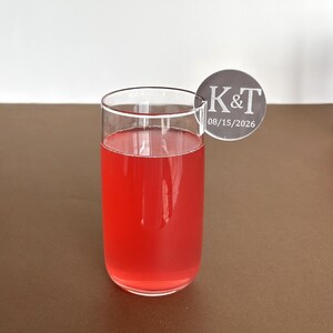 May include: A clear glass filled with a red liquid, possibly a cocktail or juice. A circular coaster with the initials "K&T" and the date "08/15/2026" is perched on the rim of the glass. The drink sits on a brown surface.
