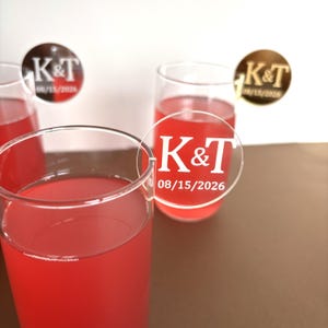May include: Clear champagne flutes filled with a red liquid, possibly a cocktail. Each glass has a clear, circular tag with the initials "K&T" and the date "08/15/2026" in white. The background is white and brown.