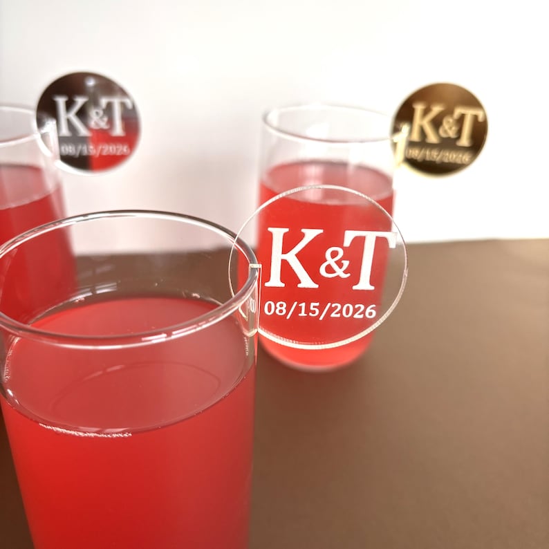 May include: Clear champagne flutes filled with a red liquid, possibly a cocktail. Each glass has a clear, circular tag with the initials "K&T" and the date "08/15/2026" in white. The background is white and brown.