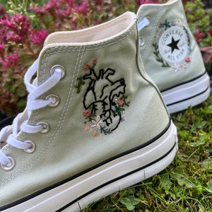 May include: Sage green high-top Converse trainers with embroidered floral details and a black heart on one shoe. The other shoe has the Converse logo embroidered with flowers.