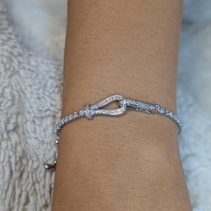 May include: A silver bracelet featuring a decorative clasp, embellished with small, sparkling stones. The bracelet is displayed on a wrist, highlighting its delicate design and intricate details. The clasp is shaped like a knot.