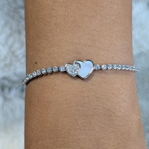 May include: A silver bracelet with a row of small, clear, sparkling stones. The bracelet has a double heart charm at the centre, one heart is encrusted with smaller stones, and the other is a smooth, pearly white. Jewellery.