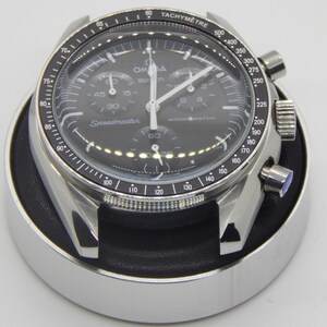 Puede incluir: Un reloj de pulsera de acero inoxidable con esfera negra, manecillas y marcadores blancos. El reloj tiene m&uacute;ltiples subesferas y el texto "Speedmaster" y "Moonwatch" es visible. El reloj est&aacute; en una caja plateada y descansa sobre un soporte negro y plateado.