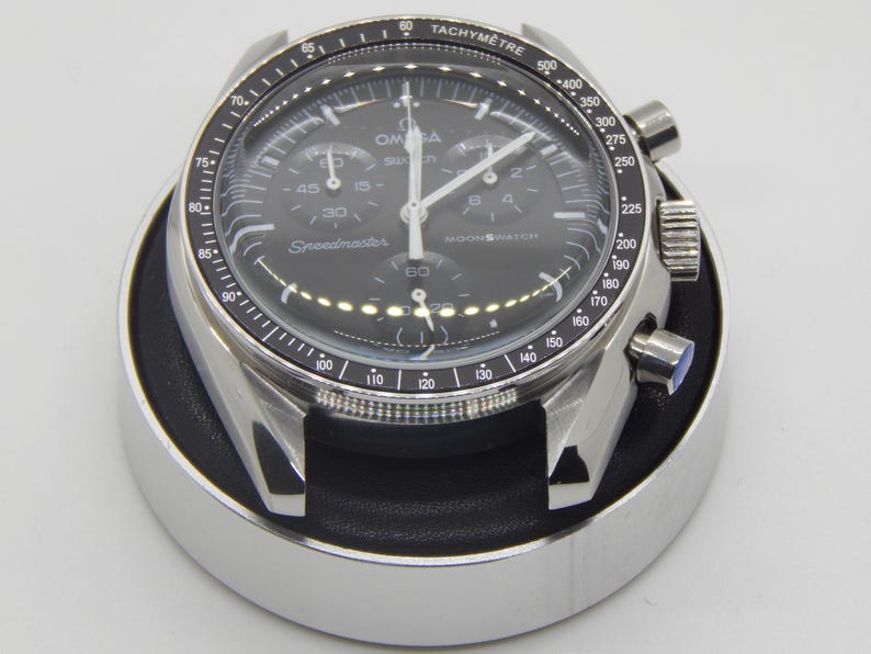 Puede incluir: Un reloj de pulsera de acero inoxidable con esfera negra, manecillas y marcadores blancos. El reloj tiene m&uacute;ltiples subesferas y el texto "Speedmaster" y "Moonwatch" es visible. El reloj est&aacute; en una caja plateada y descansa sobre un soporte negro y plateado.