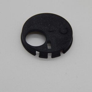 Puede incluir: Un objeto negro, circular, impreso en 3D con un gran agujero circular y una abertura rectangular m&aacute;s peque&ntilde;a. El objeto tiene una superficie texturizada y varios peque&ntilde;os recortes alrededor del borde. Se encuentra sobre una superficie blanca lisa.