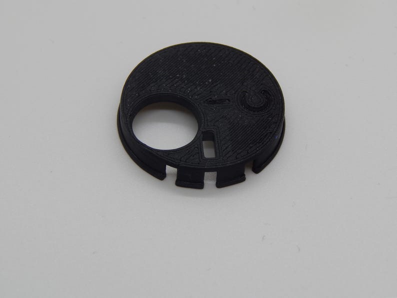 Puede incluir: Un objeto negro, circular, impreso en 3D con un gran agujero circular y una abertura rectangular m&aacute;s peque&ntilde;a. El objeto tiene una superficie texturizada y varios peque&ntilde;os recortes alrededor del borde. Se encuentra sobre una superficie blanca lisa.