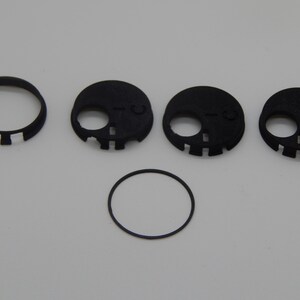 Puede incluir: Un conjunto de componentes negros impresos en 3D con forma de calavera, que incluyen un anillo y varias piezas circulares con recortes para los ojos y la boca. Los componentes est&aacute;n dispuestos sobre una superficie blanca.