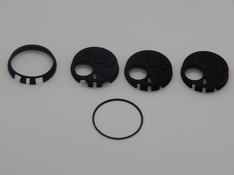 Puede incluir: Un conjunto de componentes negros impresos en 3D con forma de calavera, que incluyen un anillo y varias piezas circulares con recortes para los ojos y la boca. Los componentes est&aacute;n dispuestos sobre una superficie blanca.