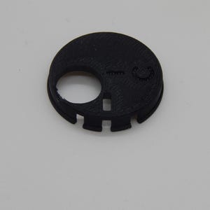 Puede incluir: Un componente de pl&aacute;stico negro, circular, con una gran abertura circular y varios recortes m&aacute;s peque&ntilde;os. El objeto tiene una superficie texturizada y parece estar impreso en 3D. El dise&ntilde;o incluye un peque&ntilde;o detalle circular en relieve y una abertura rectangular.
