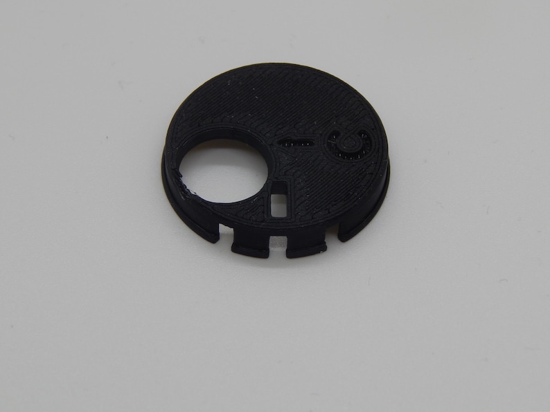 Puede incluir: Un componente de pl&aacute;stico negro, circular, con una gran abertura circular y varios recortes m&aacute;s peque&ntilde;os. El objeto tiene una superficie texturizada y parece estar impreso en 3D. El dise&ntilde;o incluye un peque&ntilde;o detalle circular en relieve y una abertura rectangular.