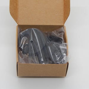 Puede incluir: Un objeto negro, posiblemente una herramienta o accesorio, est&aacute; empaquetado dentro de una peque&ntilde;a caja de cart&oacute;n abierta. El objeto est&aacute; envuelto en pl&aacute;stico transparente. La caja es rectangular y de color marr&oacute;n natural.